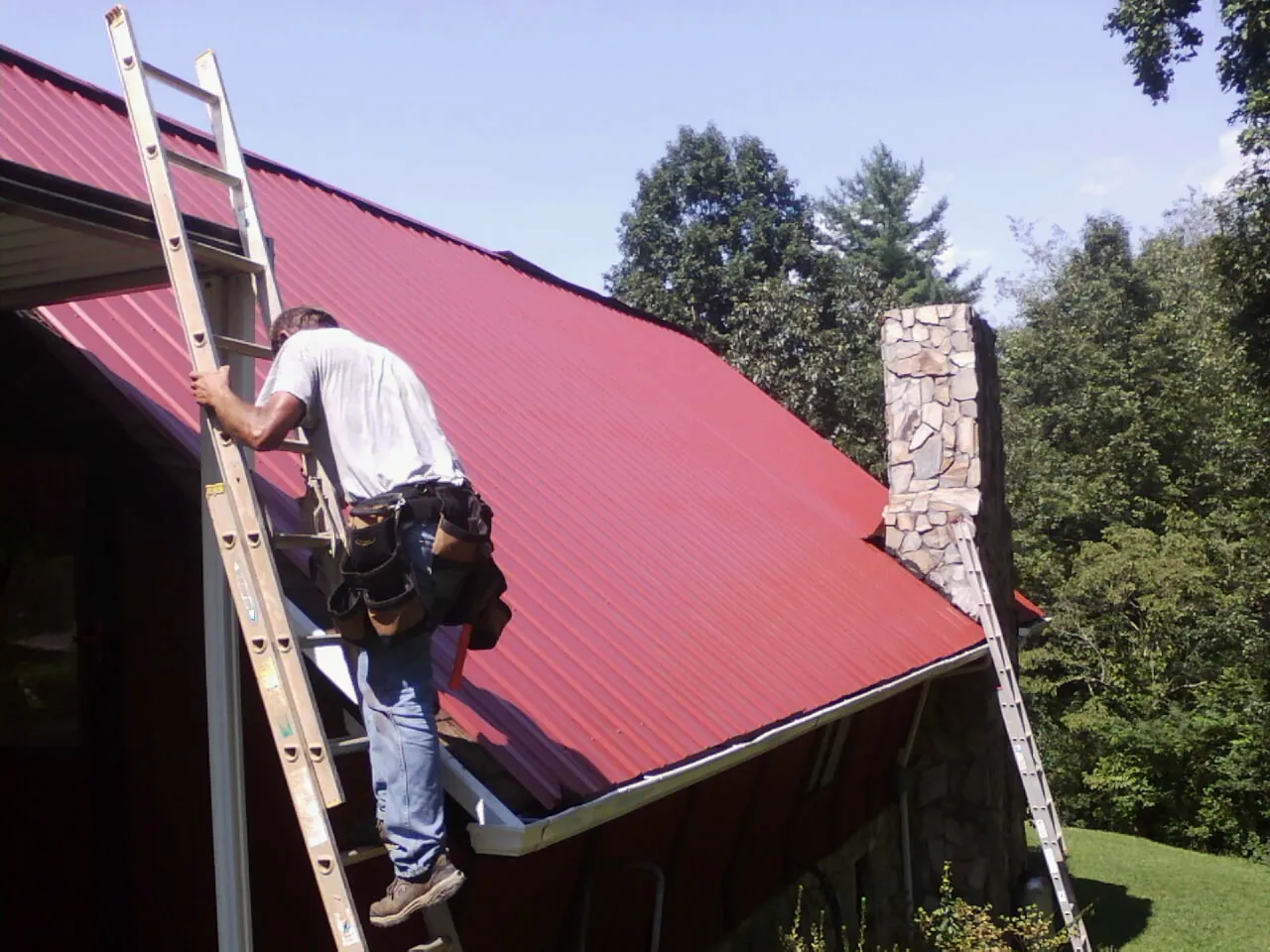 Roofer on a ladder beside a red metal roof and stone chimney — Soffit & Fascia Repair by Roofing Brothers in Wrightsboro